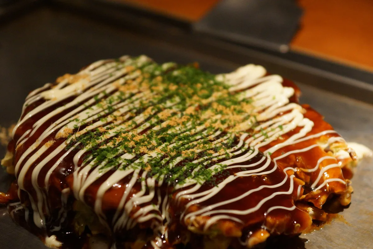 Okonomiyaki on a hot griddle, topped with brown sauce, mayonnaise and green seaweed flakes