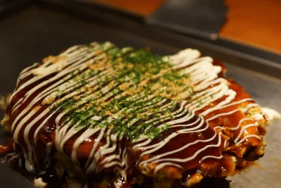 Okonomiyaki on a hot griddle, topped with brown sauce, mayonnaise and green seaweed flakes