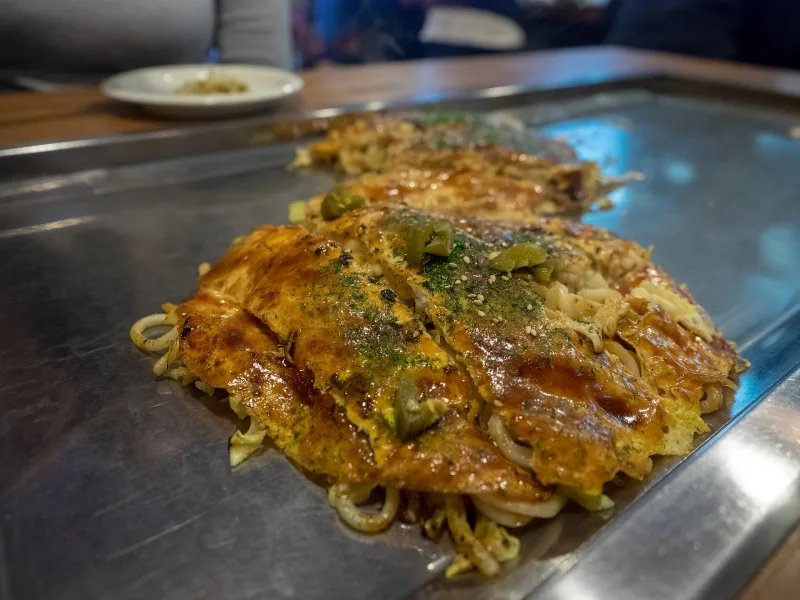 Hiroshima-style okonomiyaki on a griddle, with a layer of fried noodles visible underneath