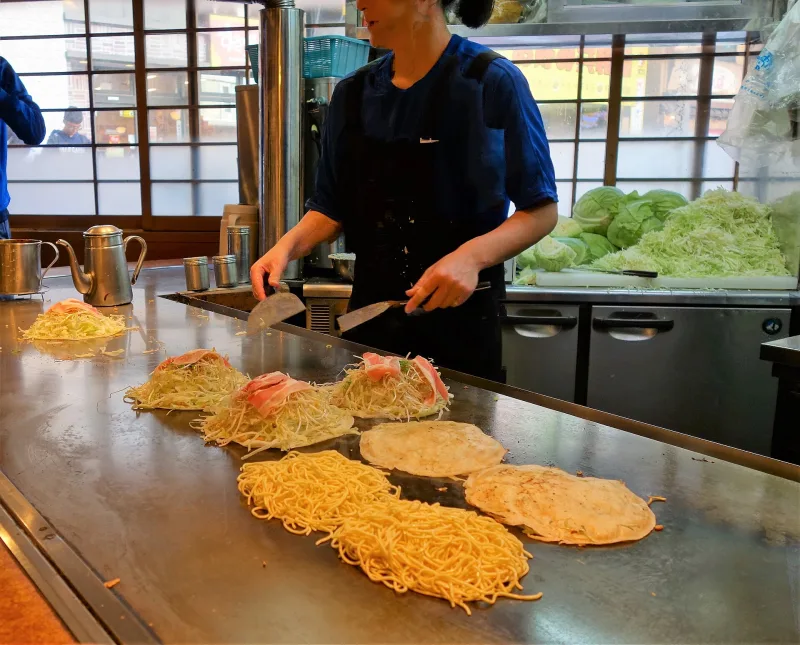Several Hiroshima-style okonomiyaki being built in layers of cabbage and noodles on a long restaurant griddle