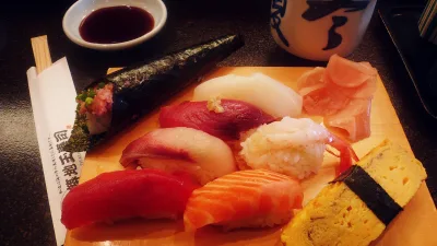 A wooden tray of assorted nigiri sushi and a hand roll, with pickled ginger and a dish of soy sauce