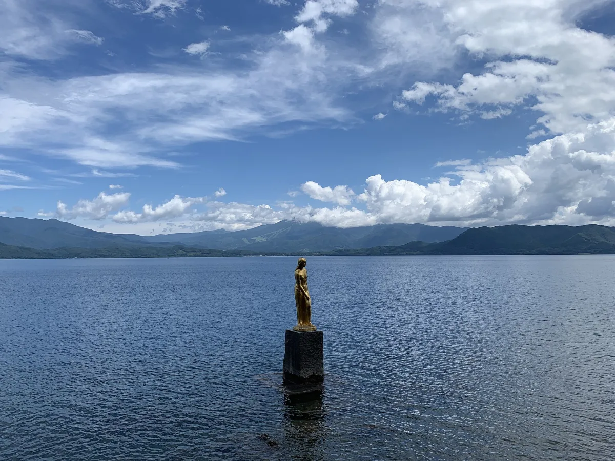 Golden Tatsuko statue standing on a black pedestal in Lake Tazawa under a cloudy sky