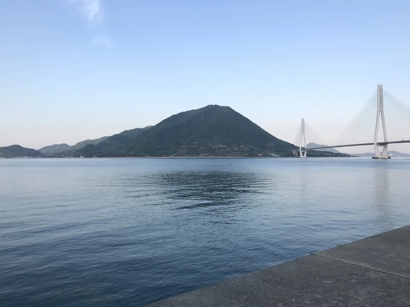 The white towers of Tatara Bridge across calm sea, next to a cone-shaped green hill