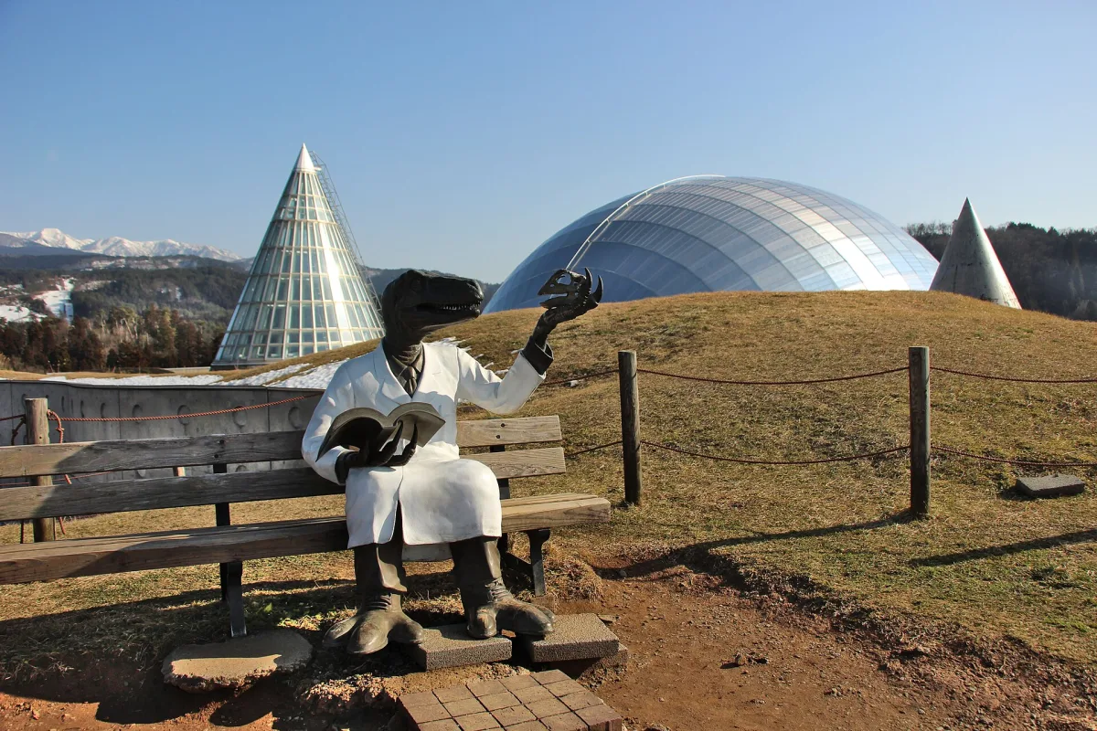 The silver dome of the Fukui Prefectural Dinosaur Museum on a hill, with a dinosaur scientist statue on a bench
