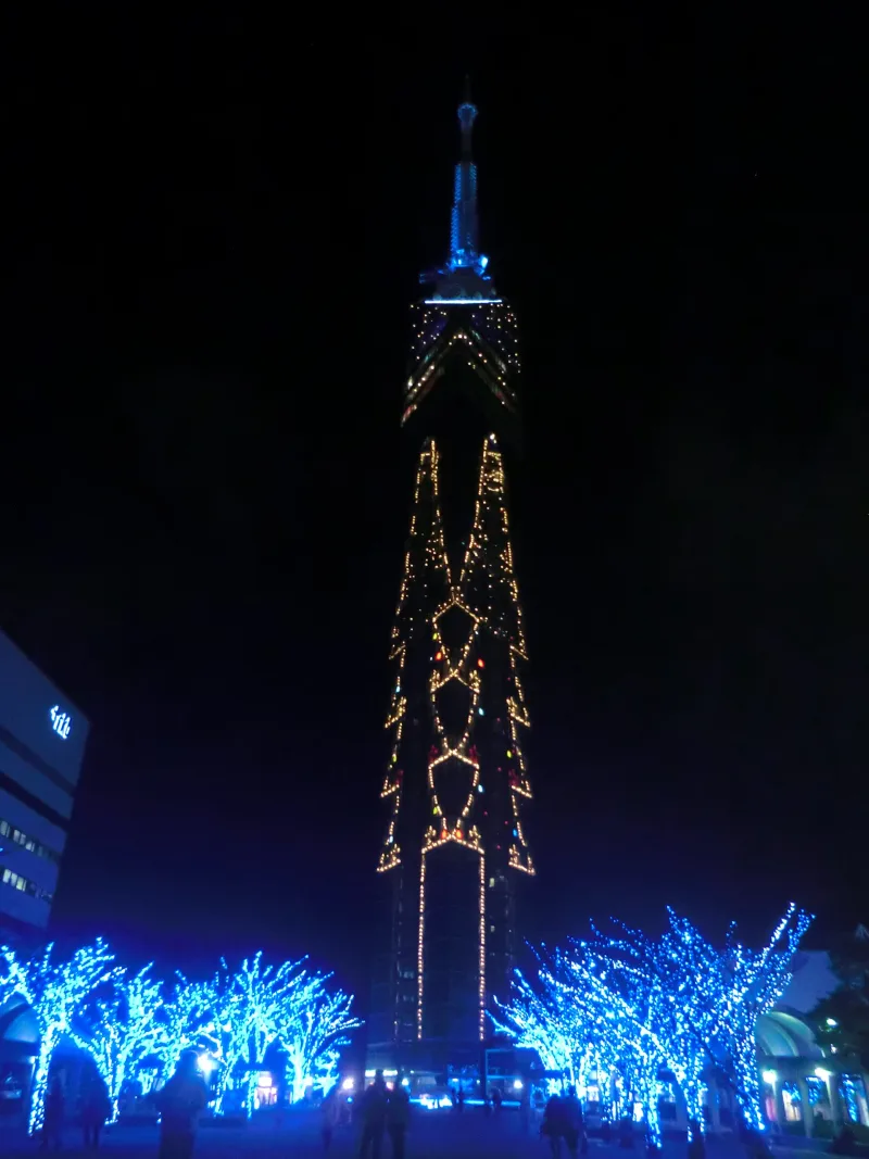 Fukuoka Tower lit with orange lights at night above trees wrapped in blue winter illuminations
