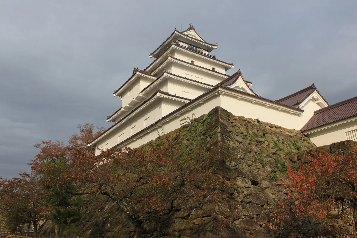 White five-story keep of Tsuruga Castle with red-tiled roofs above a stone wall and autumn trees