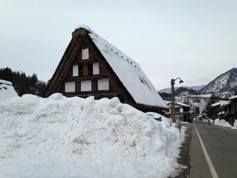 A gassho-zukuri house with a snow-covered thatched roof beside a road in winter