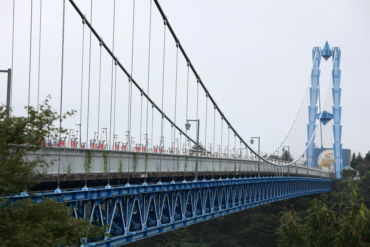 Side view of the long blue Ryujin Suspension Bridge and its tower spanning a wooded gorge
