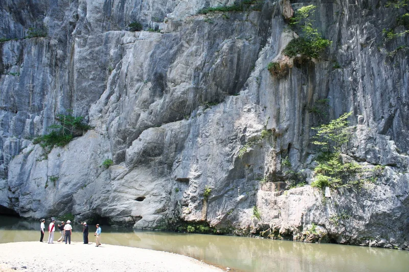 Huge pale limestone rock wall rising above a sandy riverbank, with visitors at its foot