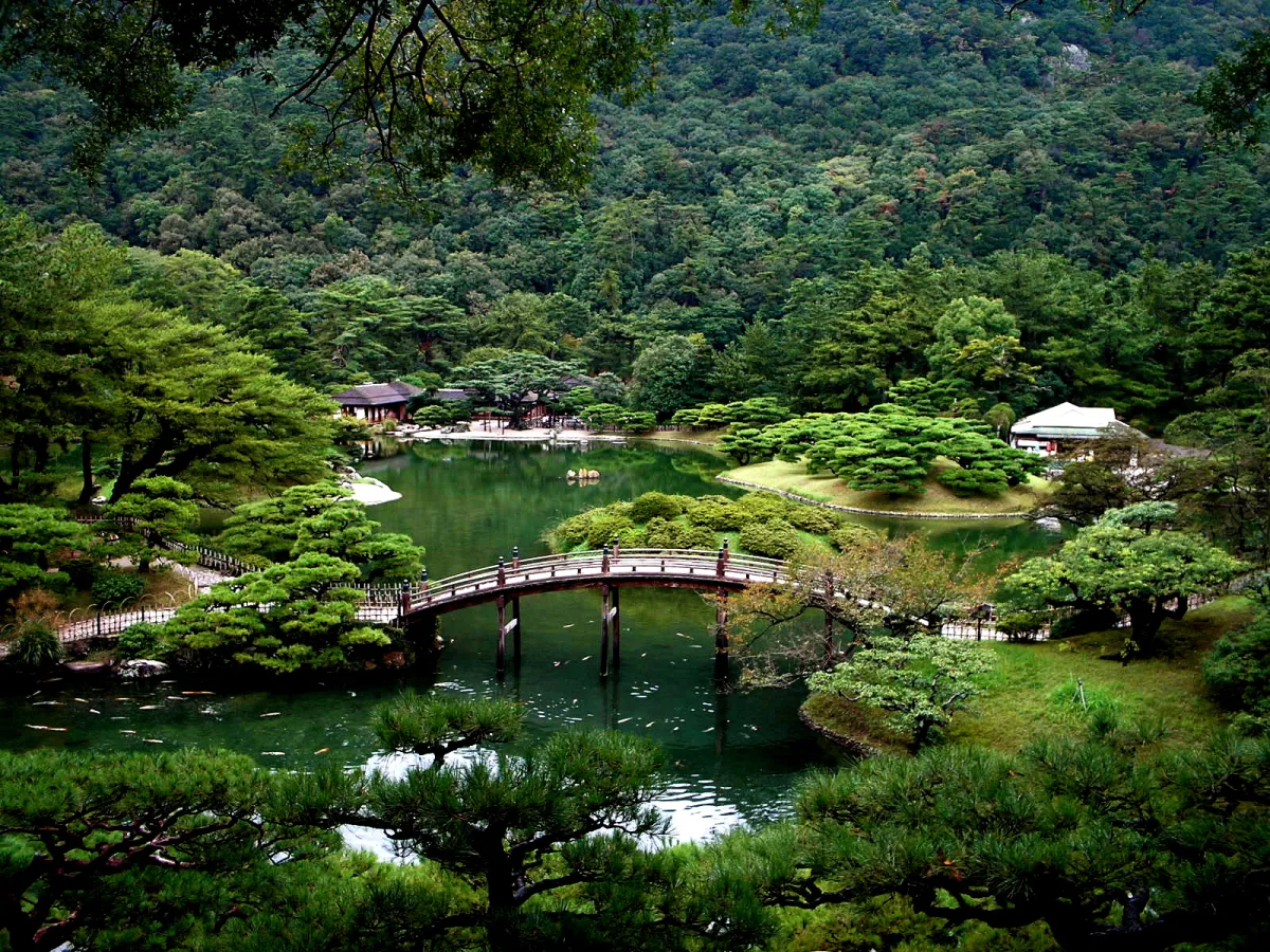 An arched bridge over a green pond framed by carefully pruned pines in Ritsurin Garden