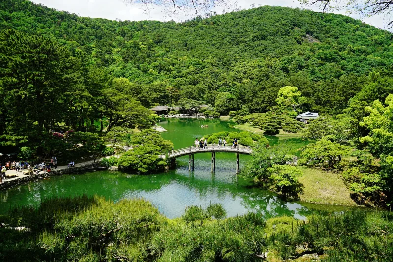 A pond with a small arched bridge below the wooded slopes of Mount Shiun at Ritsurin Garden