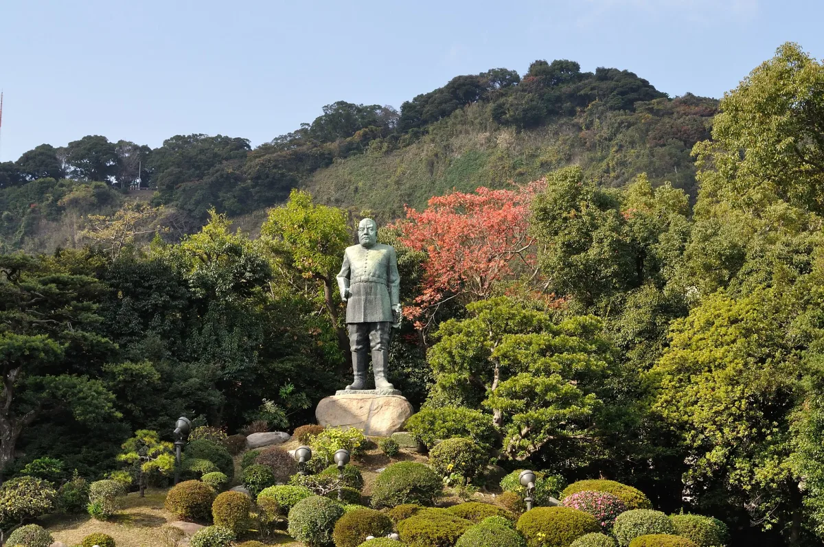 Bronze statue of Saigo Takamori in army uniform standing in a garden with Shiroyama Hill behind