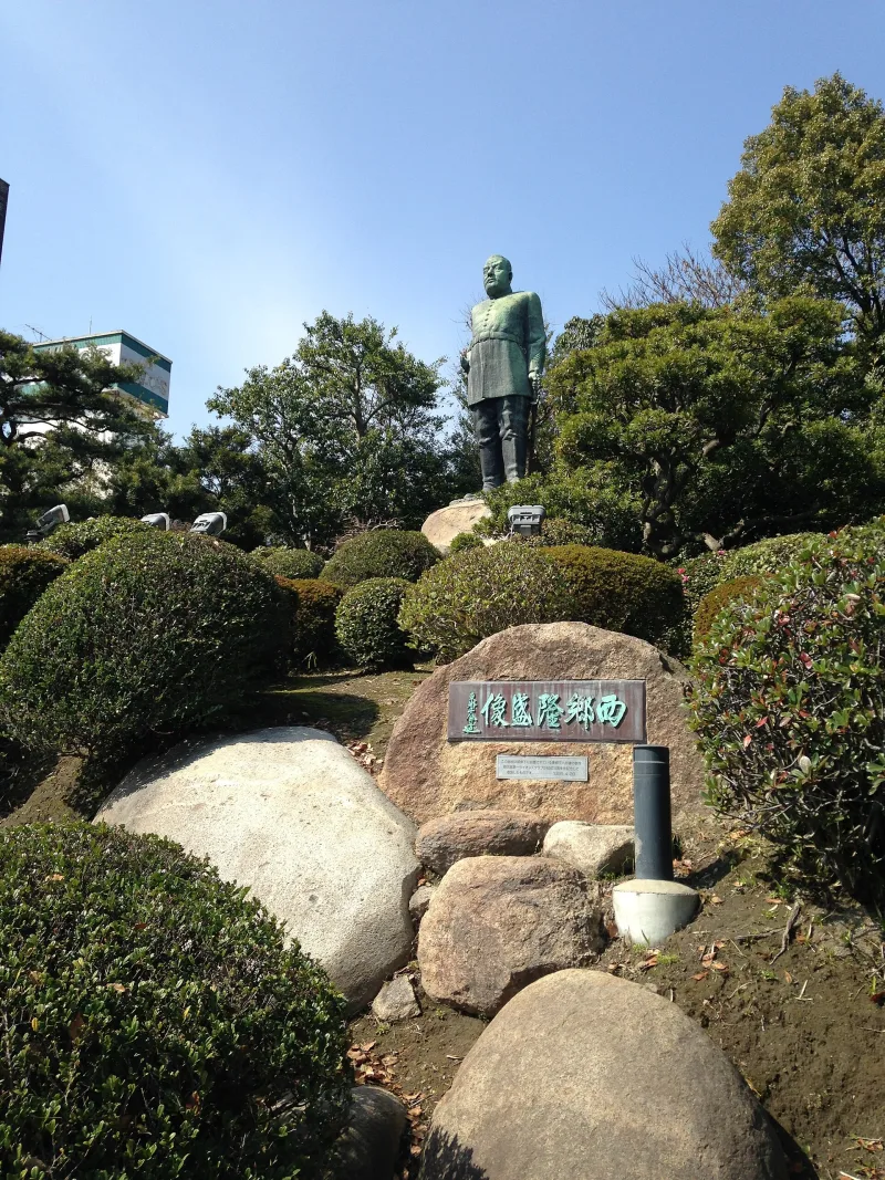 The Saigo Takamori statue in Kagoshima seen beyond landscaped shrubs and a stone nameplate