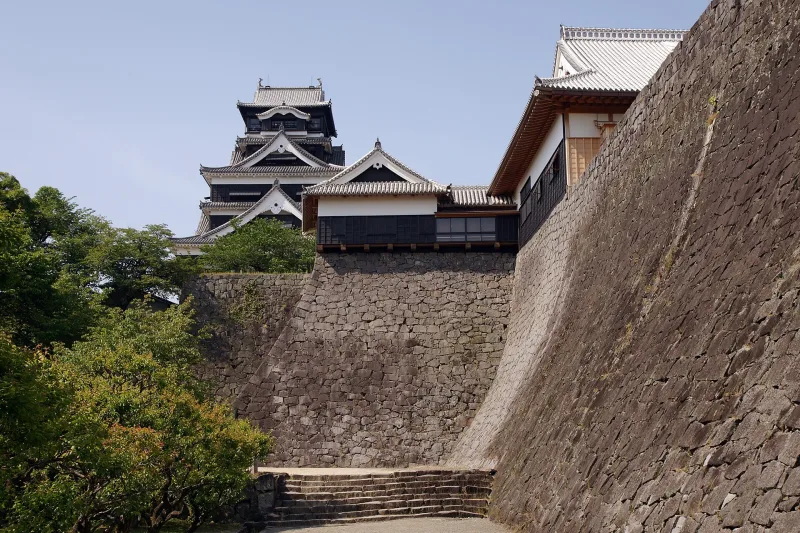 Massive curved stone walls and a turret of Kumamoto Castle with visitors below