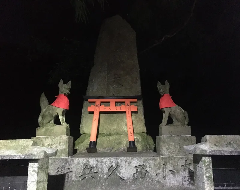 A pair of stone fox statues in red bibs beside a small torii at night