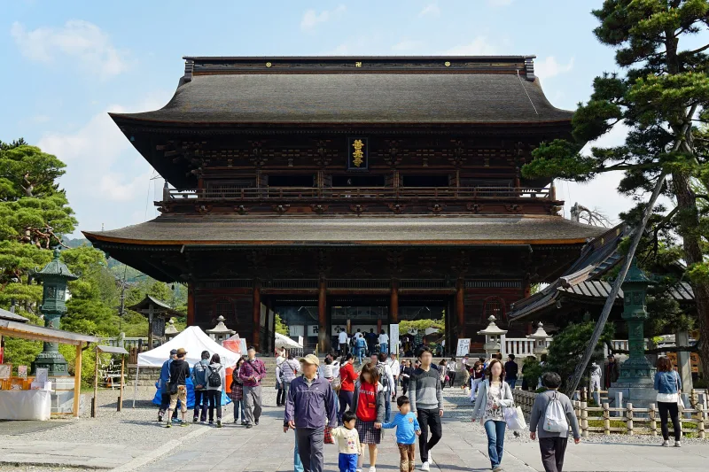 The two-storey Sanmon gate of Zenko-ji seen from the approach, with visitors walking toward it
