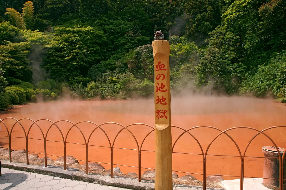 Steam rising from the orange-red pond behind a low fence at Chinoike Jigoku in Beppu