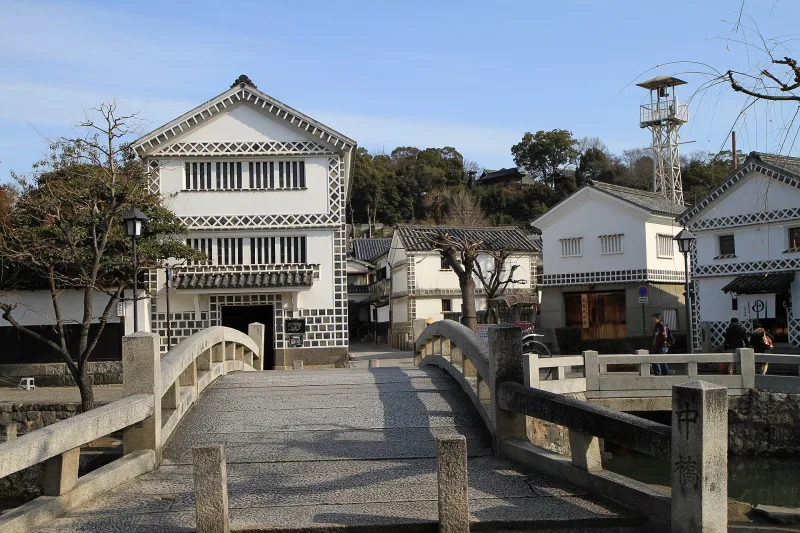 White-walled storehouse with black-and-white namako tile walls beside a stone bridge in Kurashiki