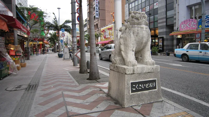 Stone shisa lion statue marking Kokusai-dori on a palm-lined sidewalk in Naha