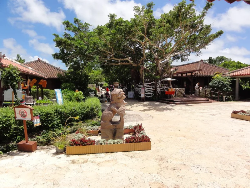 Plaza in Okinawa World with red-tiled buildings, trees and a shisa lion statue