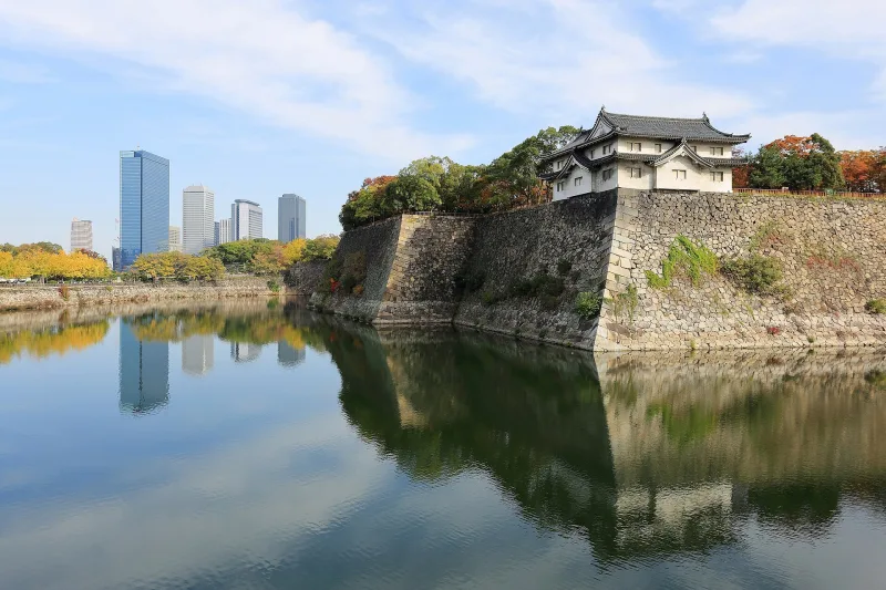 Inui-yagura turret on a stone wall reflected in Osaka Castle's outer moat, office towers behind