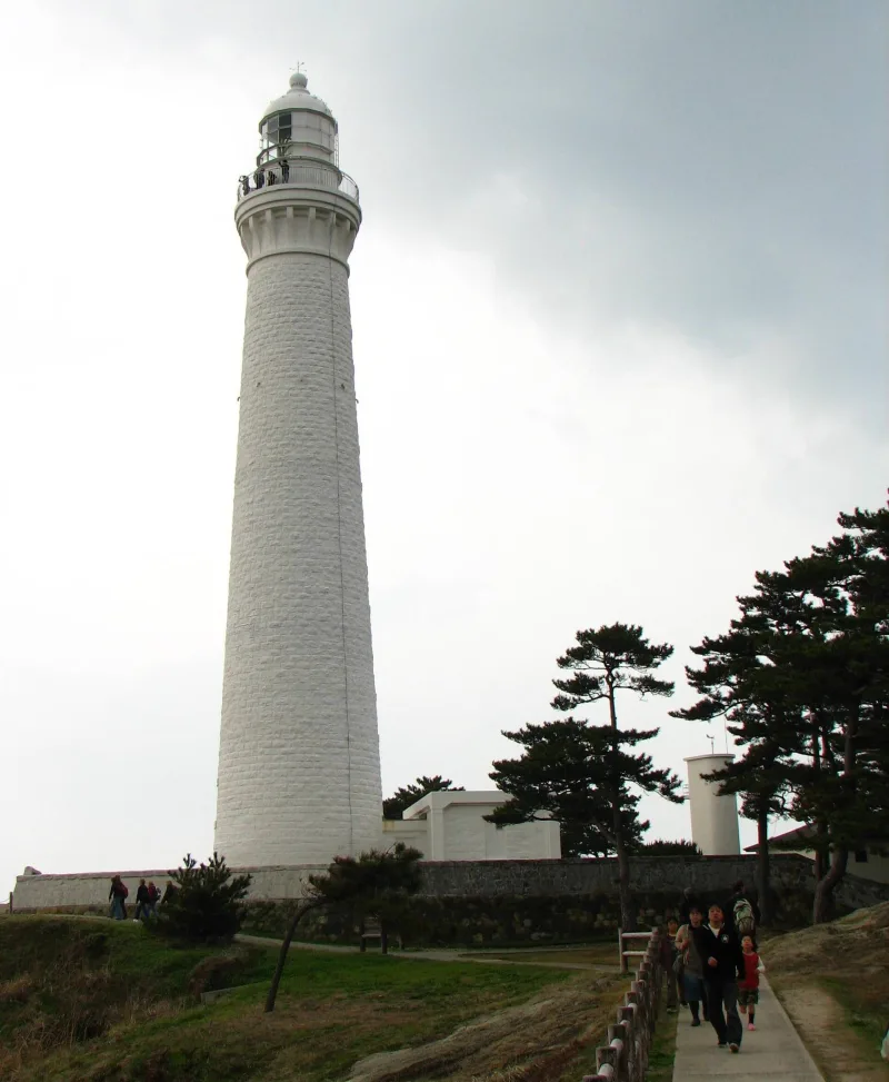 Visitors walking toward the tall white tower of Hinomisaki Lighthouse among pine trees