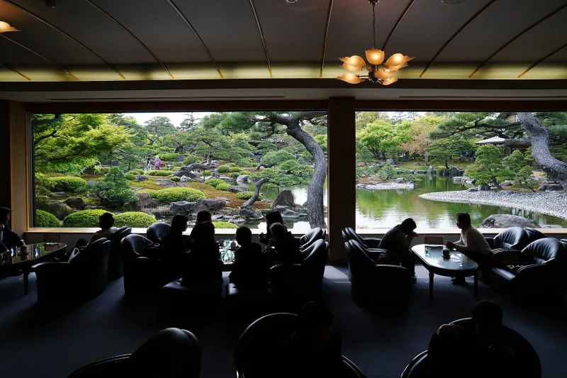 Visitors in a dim tearoom looking out through wide windows onto the Yuushien garden