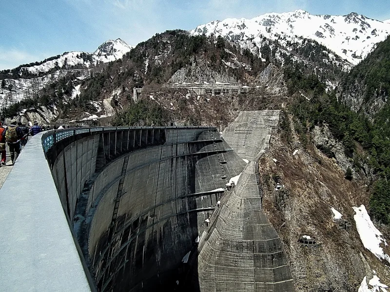 The curved crest of Kurobe Dam with snow-capped Northern Alps peaks in spring