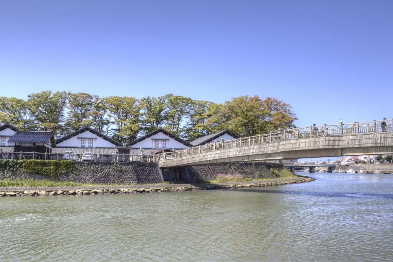 Sankyo warehouses and trees seen across the river with a footbridge in Sakata