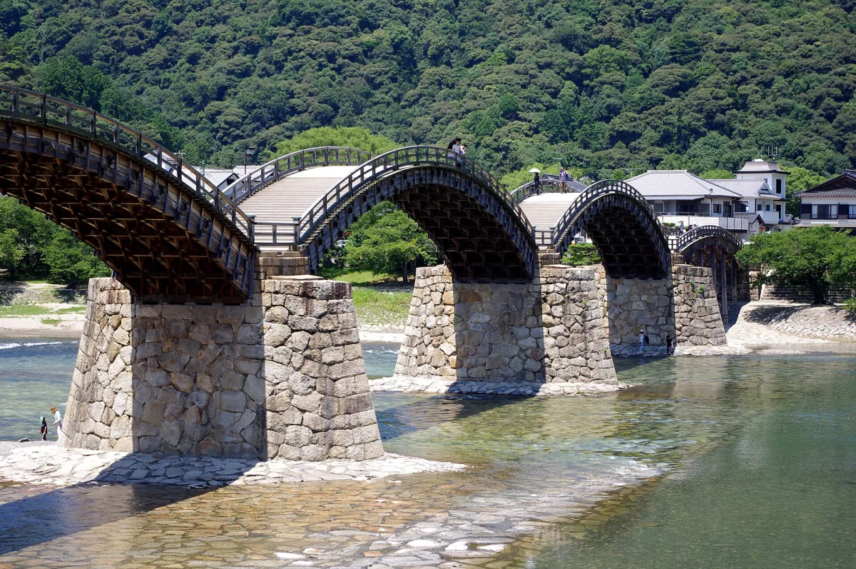 The wooden arches of Kintaikyo Bridge on stone piers over the clear Nishiki River, with green hills behind
