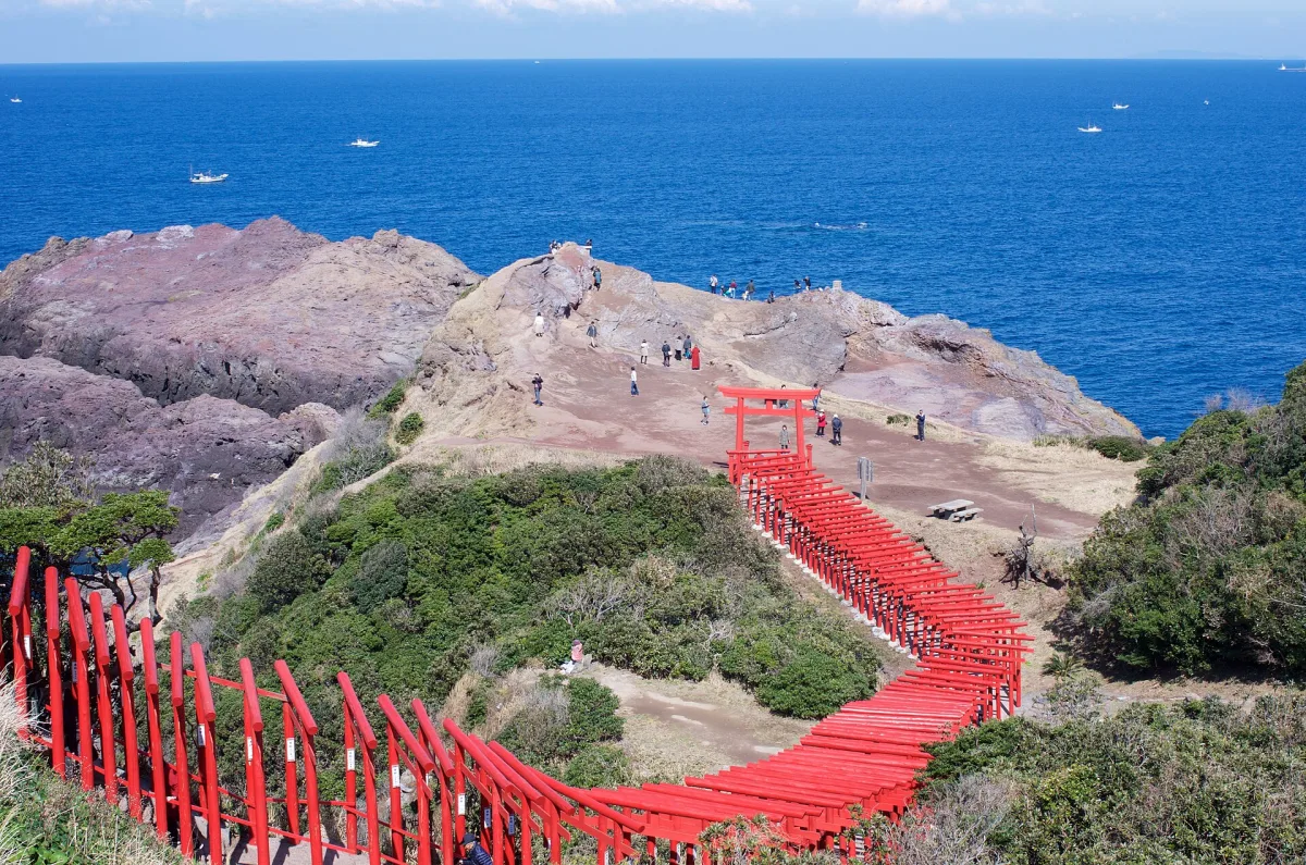 A long line of red torii gates winding down a green cliff toward the deep blue Sea of Japan at Motonosumi Shrine