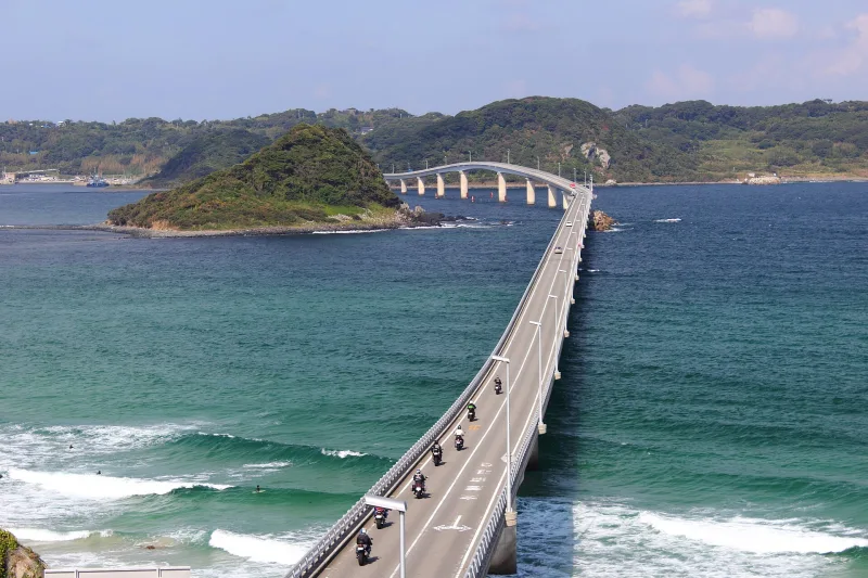 Tsunoshima Bridge running straight over green-blue water past a small wooded islet